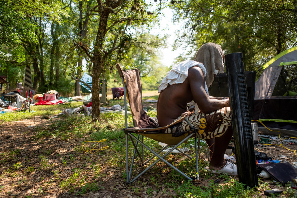 Someone shirtless with a towel draped over their head sits in a folding chair outdoors beneath a canopy of trees near a tent and various household items, including a tower-style fan and yellow extension cord.