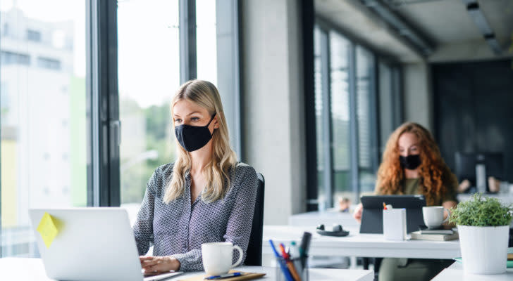 two women working on laptops in office space with black PPE face masks on