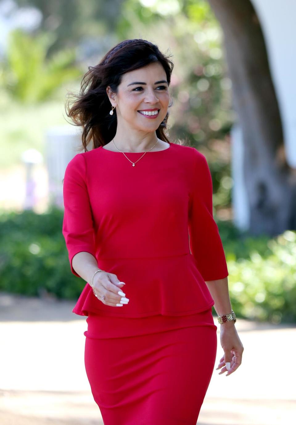 A smiling woman in a red suit walks toward the camera outdoors.