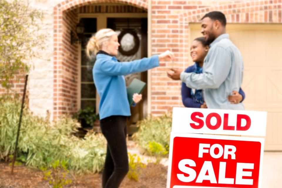 A person hands a key to another person in front of a house with a for sale sign in the lawn.