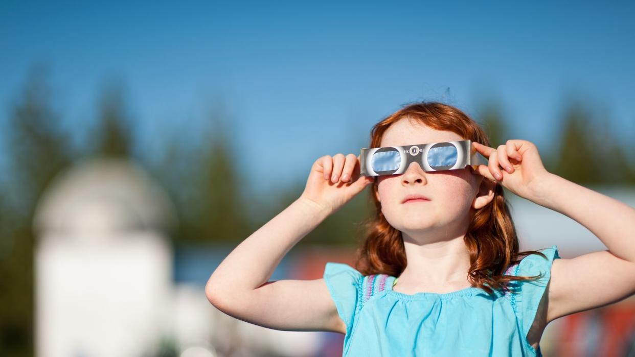 young red headed girl watching solar eclipse safely with i s o 1 2 3 1 2 2 filter glasses, silo in the background