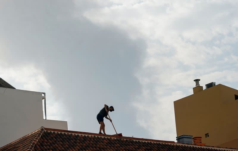 A person cleans the ash from a roof as smoke rises in the background following the eruption of a volcano on the Canary Island of La Palma