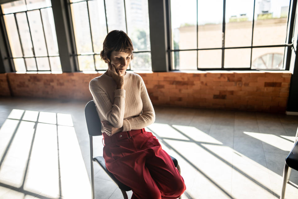 A person with short hair, wearing a long-sleeved top and red pants, sits thoughtfully on a chair in a sunlit room with large windows