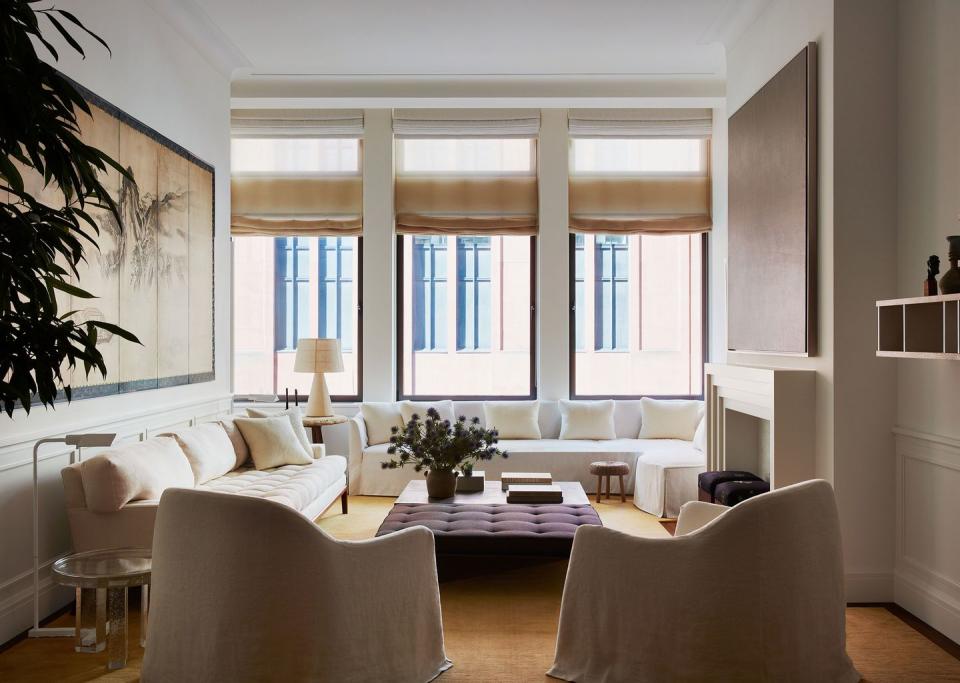 a living room in beige colors with three large windows at the back and sofas and chairs creating a square seating area with a large purple tufted ottoman at center