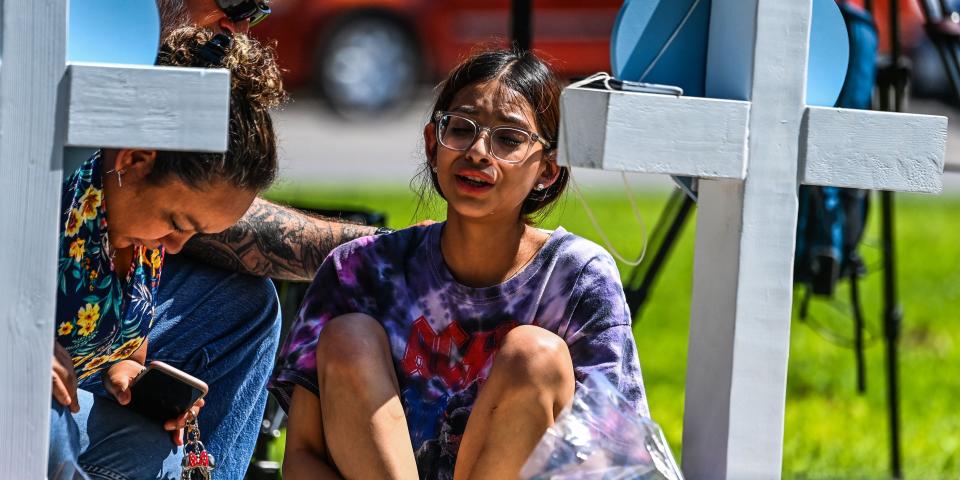 A girl sits in front of a cross holding her knees and crying.