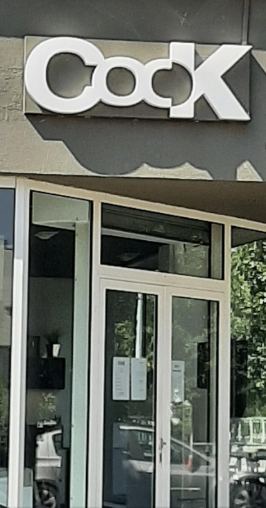 Storefront with a large sign that reads "Cook" above the entrance. The store has large windows and glass doors, with visible reflections