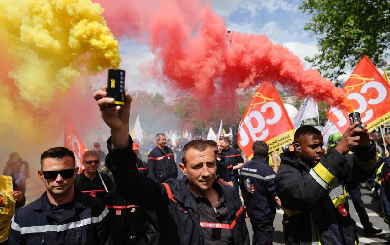 Firefighters in their uniforms marching in the sunshine while holding up smoke bombs, emitting colourful plumes