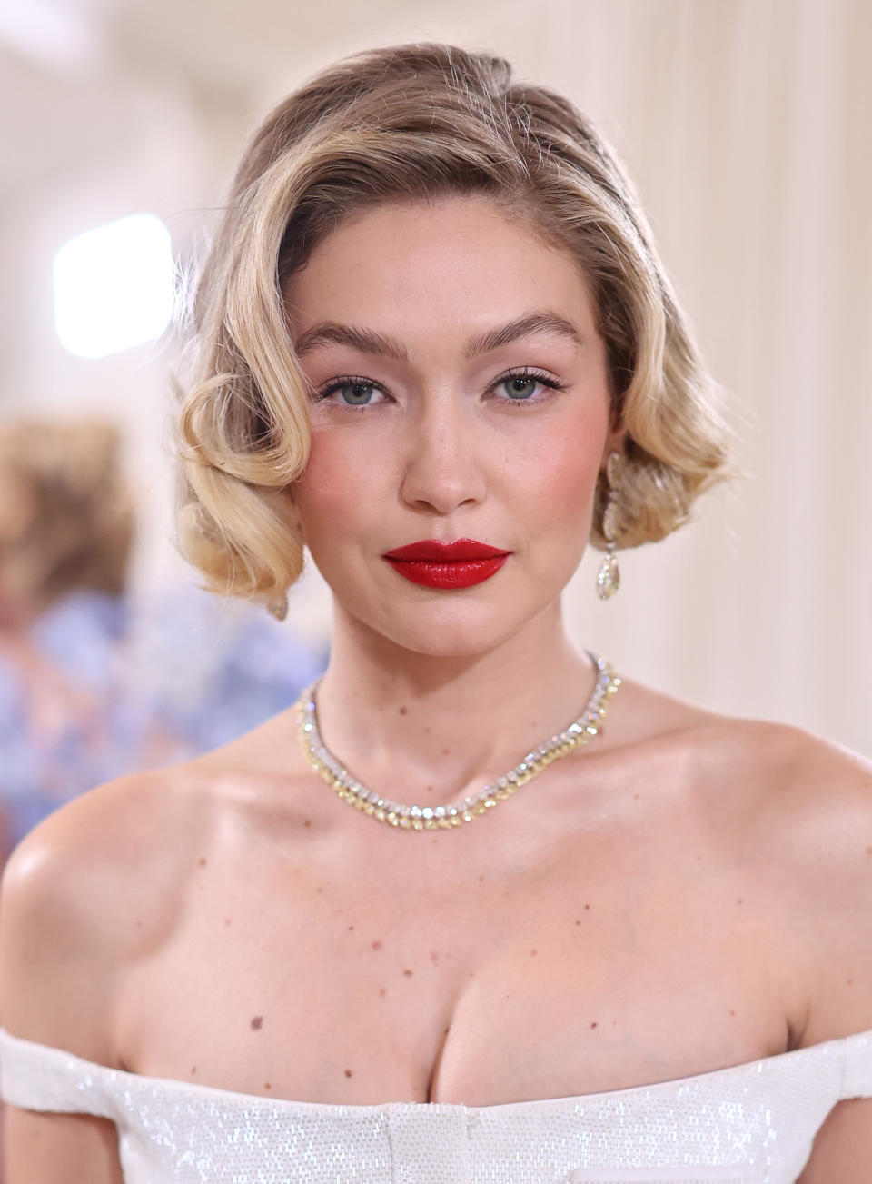Gigi Hadid is pictured in an off-the-shoulder dress with a sparkling necklace, short wavy hairstyle, and bold red lipstick