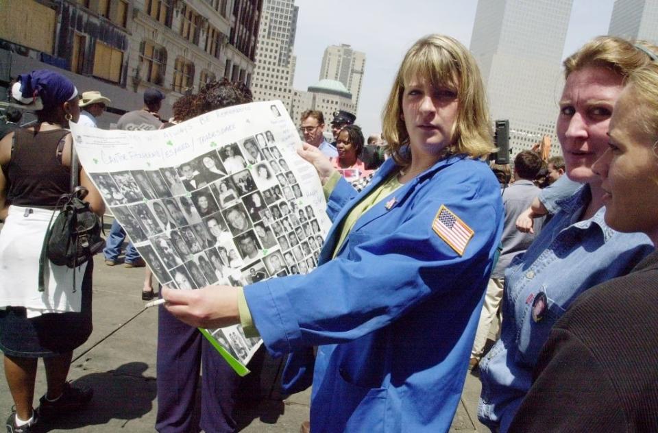 A woman wearing a blue jacket with an American flag patch holds a poster showing numerous black-and-white photos in a crowd. Buildings are in the background