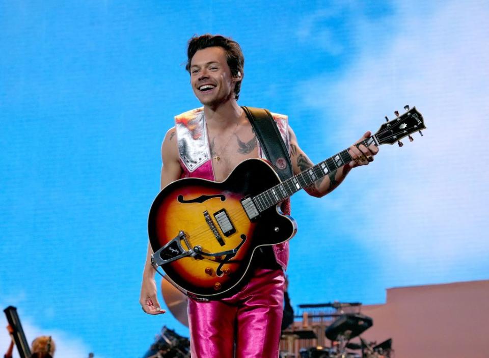 harry styles smiles while holding an electric guitar and standing on a stage