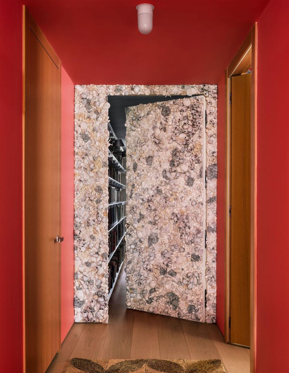 in a hallway with deep rose colored walls are two facing wood doors and at the end of the hall is a door encrusted with seashells that is open just a crack to reveal a room with shelves along the wall