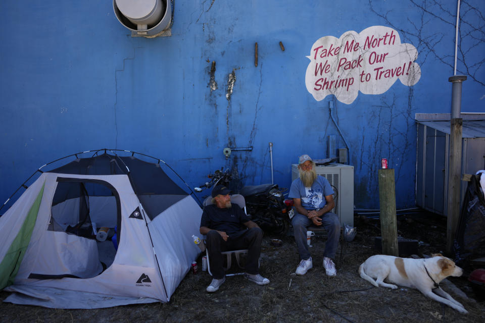 Shrimp boat workers Shawn Shelton, left, and Doug Fundak relax next to the tent where they are living since the boat they worked on was grounded and Shelton's trailer was destroyed, at Erickson & Jensen Seafood on San Carlos Island in Fort Myers Beach, Fla., Friday, Oct. 7, 2022. The pair, along with Shelton's dog Lucky, rode the storm out on "Night Wind," as surge waters and wind carried it onshore and then into an apartment building. After the storm, many in the local shrimp industry find themselves not only out of work, but also homeless, with most of the boats where they lived aboard left stranded on dry ground or heavily damaged.(AP Photo/Rebecca Blackwell)