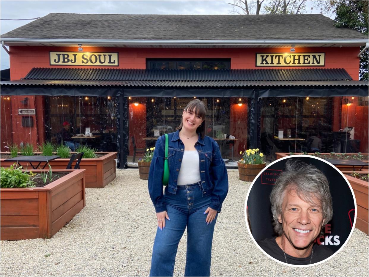 A girl smiling in font of a dark red and black awning restaurant with "JBJ Soul Kitchen" signs and a beige stone ground covered in wooden planters with plants. Jon Bon Jovi's face is blown up in a circle.