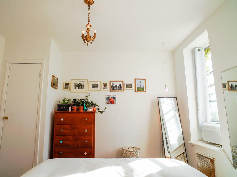 The author's bedroom in Brooklyn has white walls and a bed with a white blanket, a dresser on the left, and a mirror on the right next to a window.