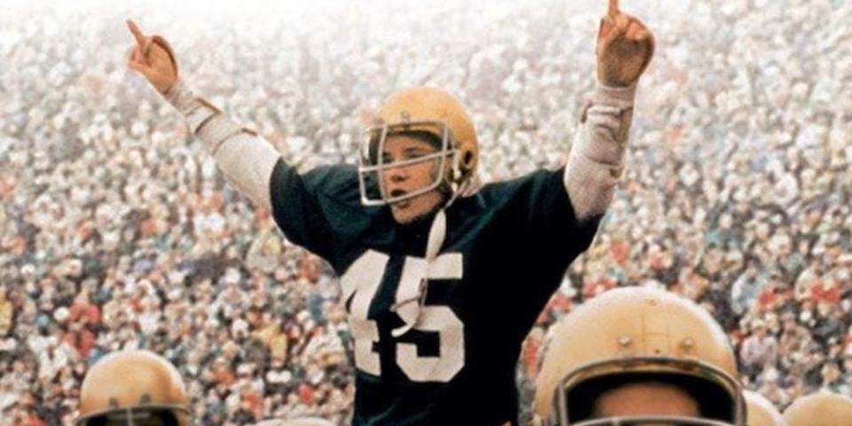 Sean Astin with his hands raised while wearing a football jersey and helmet