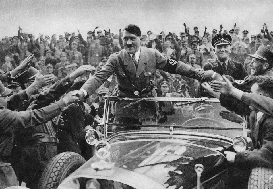 Adolf Hitler, standing up in an open car, shakes the hands of supporters.
