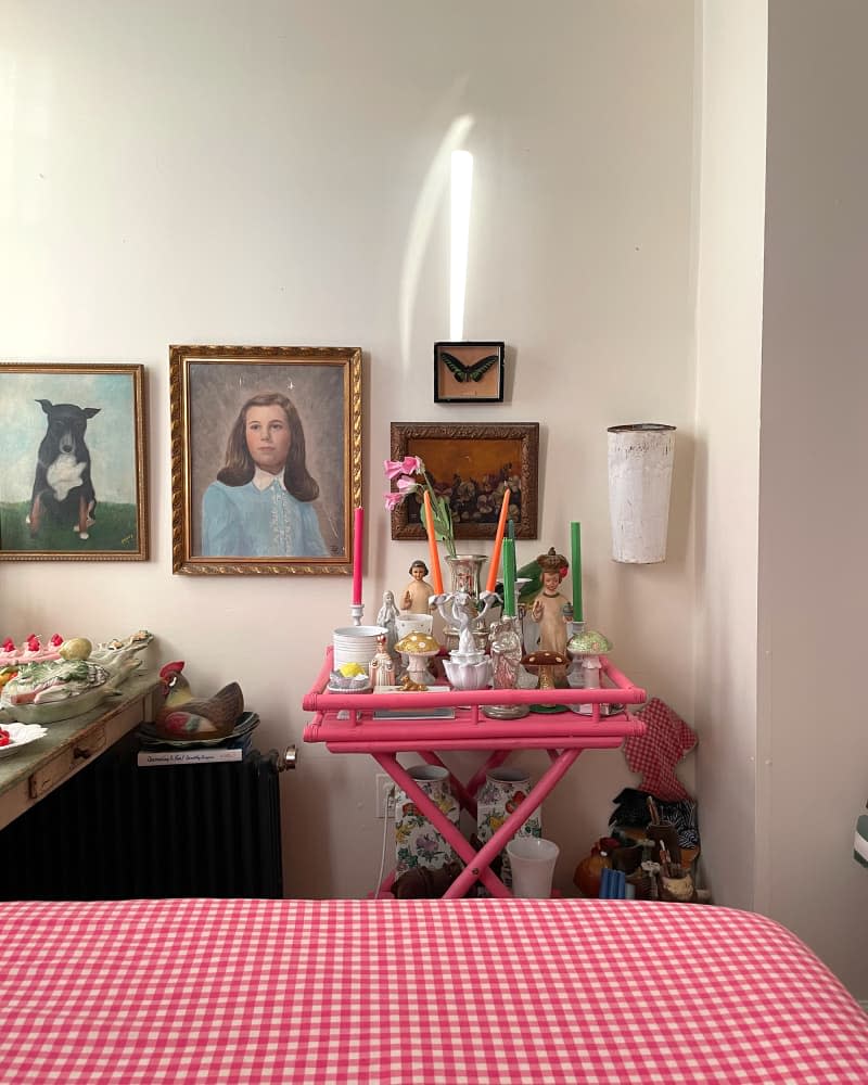 Art on white wall above pink cart with religious and mushroom decor and candlesticks in bedroom.