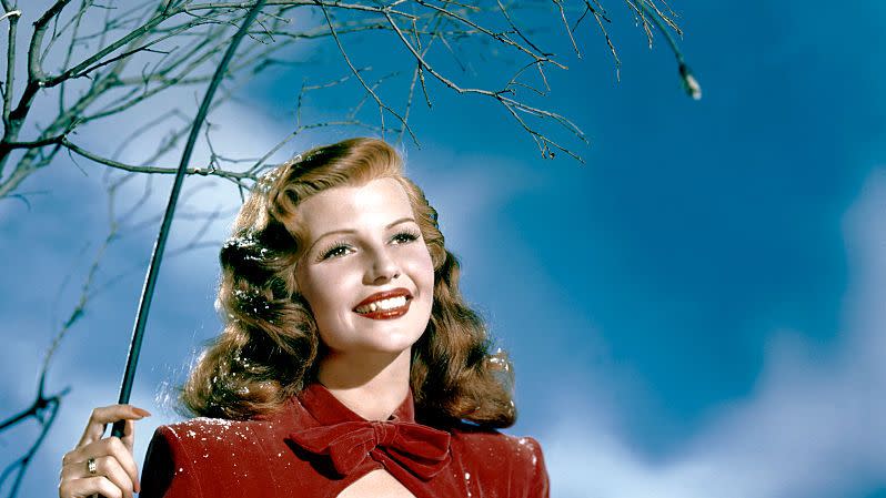 rita hayworth sits and smiles into the distance, she wears a red velvet dress with fur trim