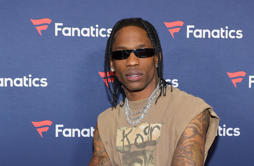 Travis Scott at a Fanatics event wearing sunglasses, a sleeveless shirt, and layered necklaces