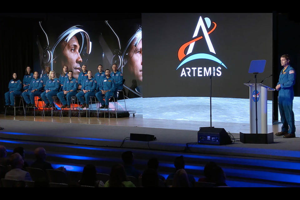 a man in a blue flight suit stands at a lectern on a stage.  behind him and to his left is a screen showing the moon's surface, the word 