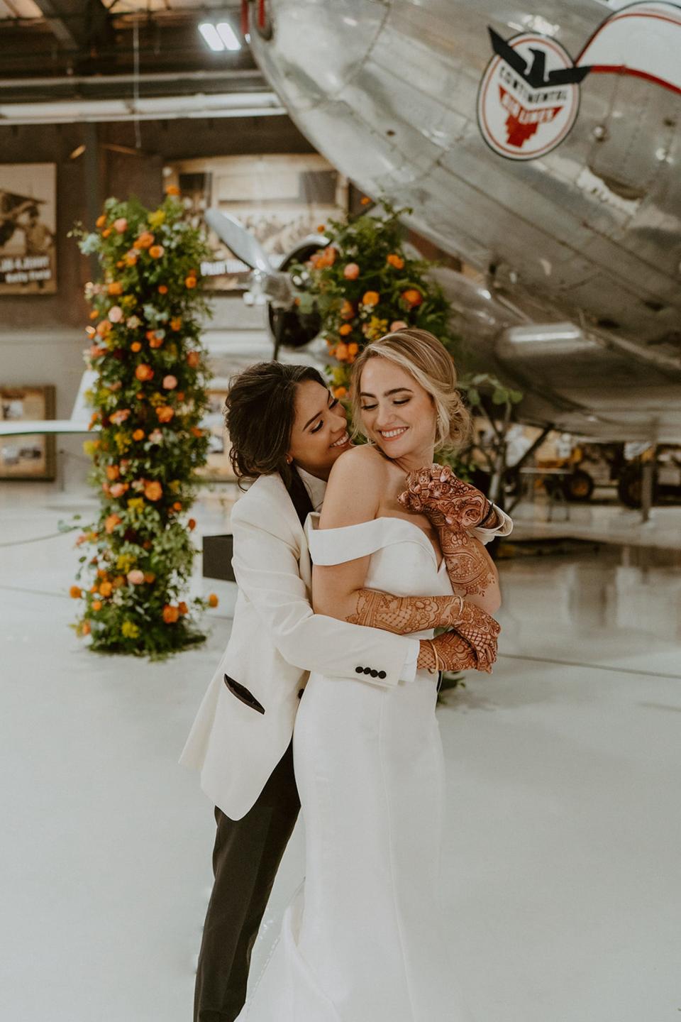 A bride hugs her wife from behind in front of a floral archway and plane.