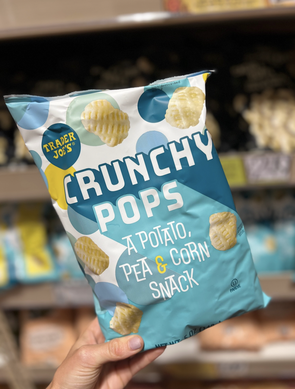 A hand holds a bag of Trader Joe's Crunchy Pops, a potato, pea, and corn snack, in a grocery store aisle