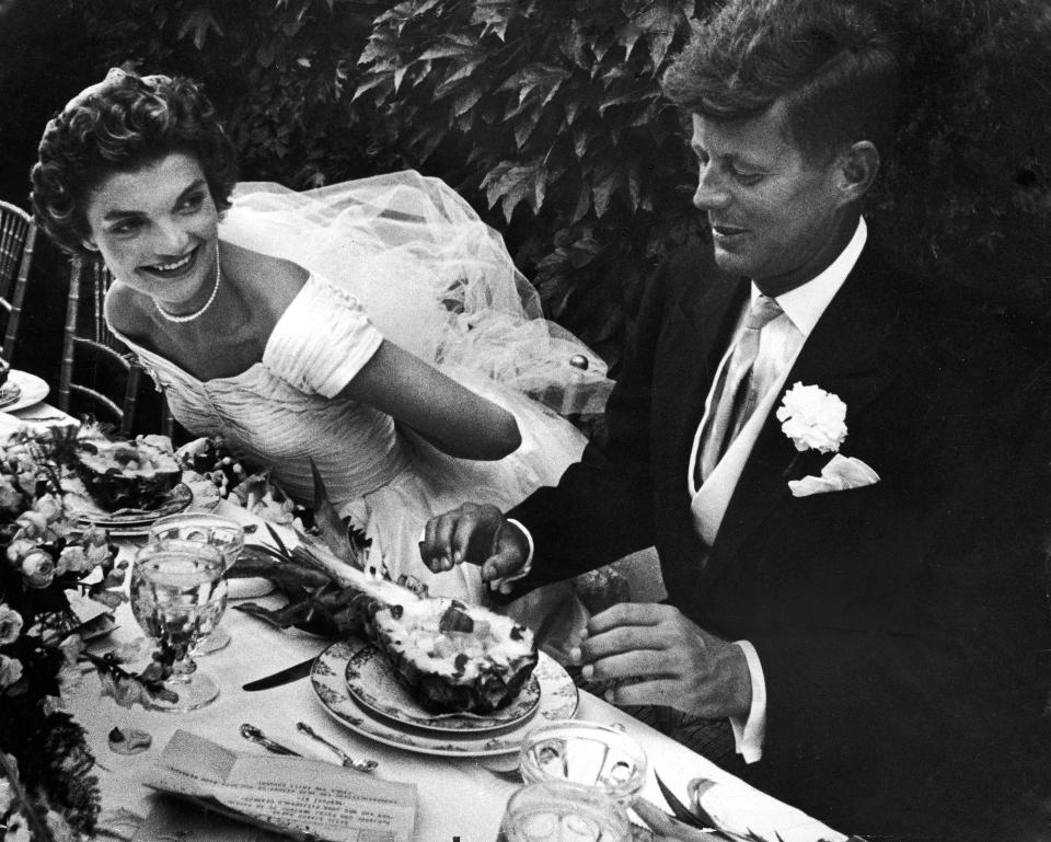 RHODE ISLAND, UNITED STATES - 1953: Senator John Kennedy & his bride Jacqueline in their wedding attire, as they sit down together at table to begin eating a pineapple salad at formally set table outdoors at their wedding reception. (Photo by Lisa Larsen/The LIFE Picture Collection/Getty Images)