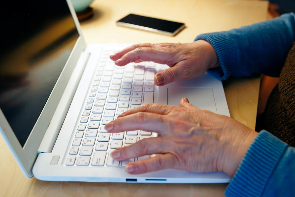 Close-up view of an elderly woman's hands typing on a laptop at home and a mobile phone lying next to her
