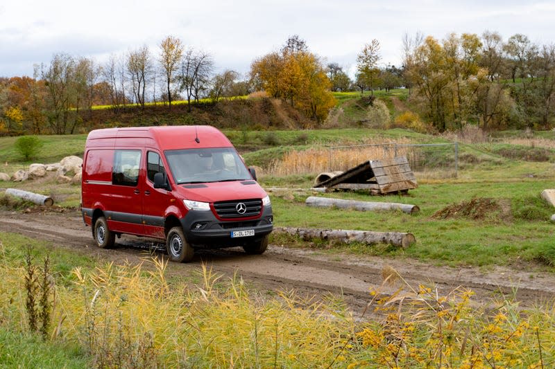 a red 2023 mercedes benz sprinter awd van driving on a dirt trail through a field, with autumn trees and rolling hills in the background