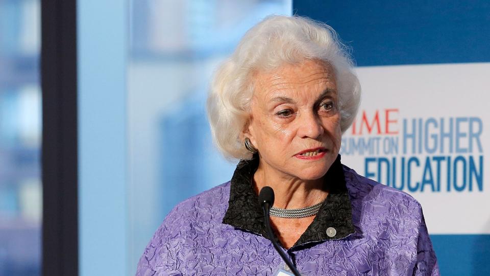 sandra day oconnor stands at a podium with a microphone and speaks, she wears a purple jacket, silver necklace and earrings