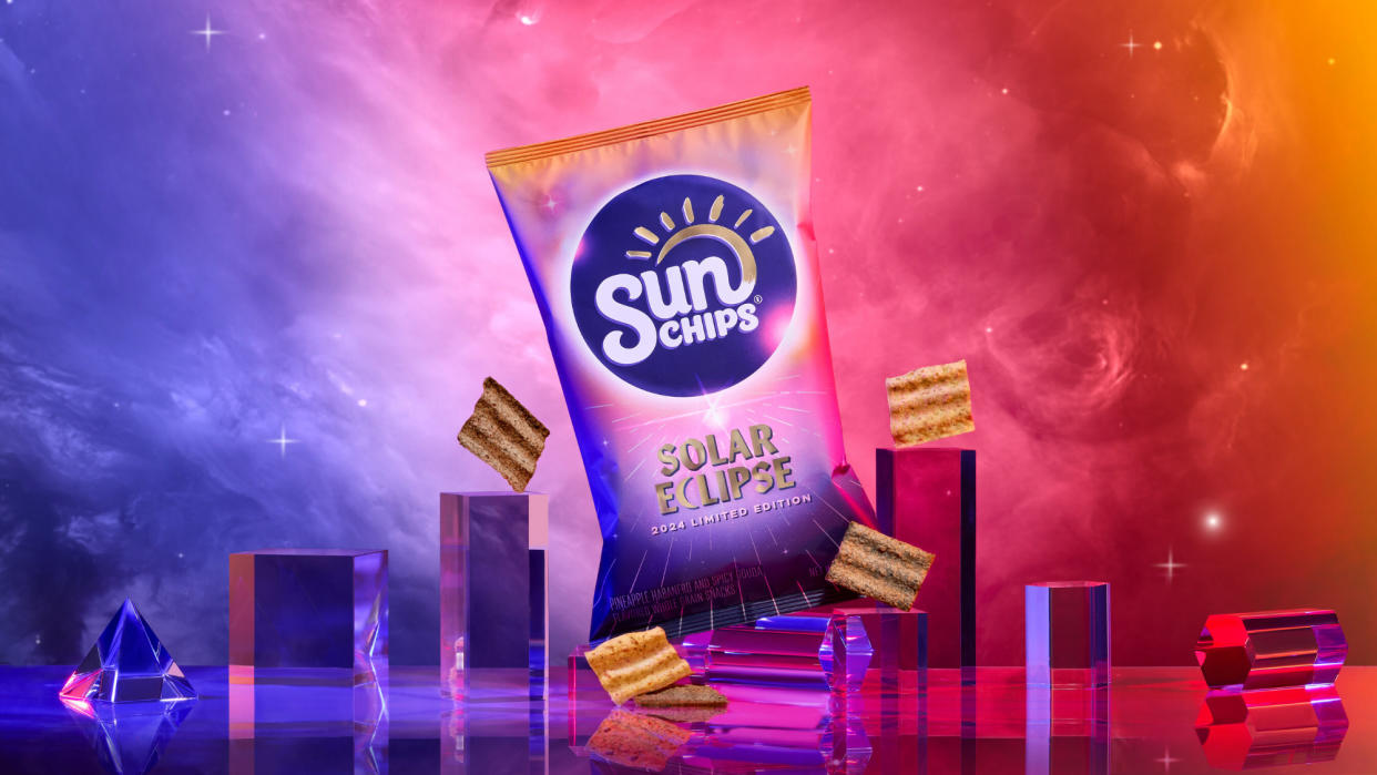  Spacey gases of purple pink red and orange collide in the background. A similarly colored back of SunChips stands in the middle. some chips float nearby. the bag is flanked on either side by transparent rectangular columns. 