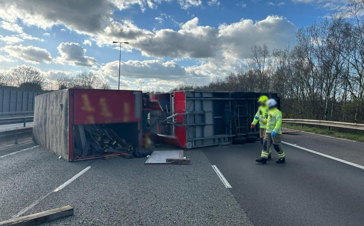 Two workers in hi-vis jackets walk across a motorway with an overturned lorry behind them