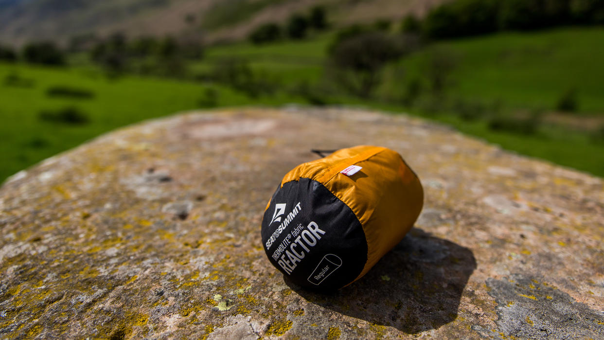  A Sea to Summit Reactor sleeping bag liner, rolled up in its bag, on a rock with fields in the background. 