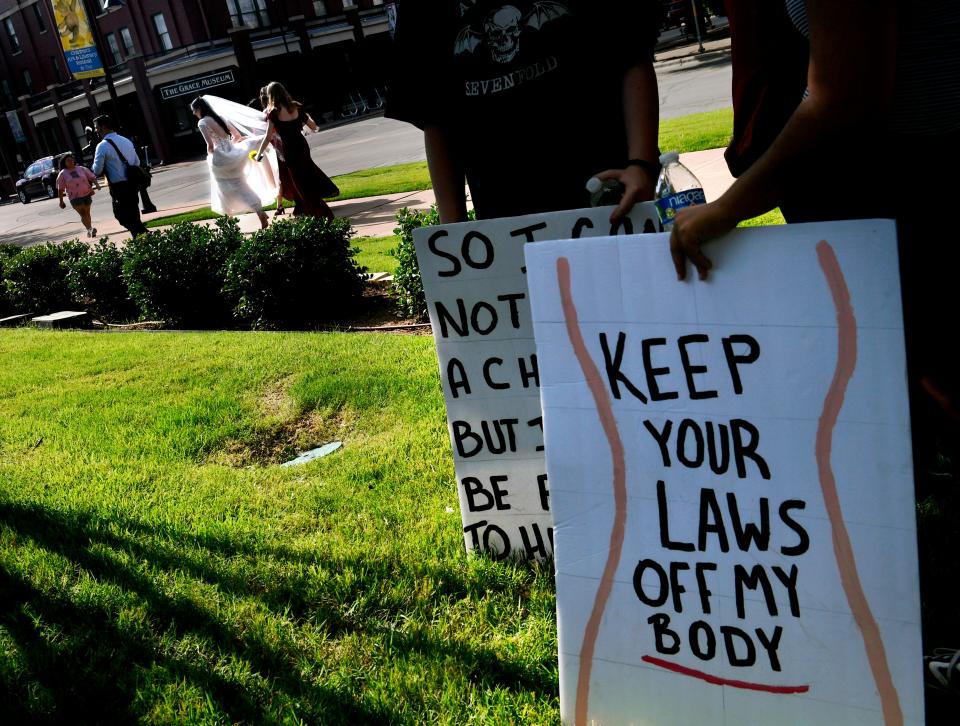 Rally participants hold signs and wait to march as Melissa Horton walks by in the background wearing her wedding gown Saturday. The bridal party and abortion rights rally amicably shared the space, enabling the bride and groom to make their pictures in the park without incident.