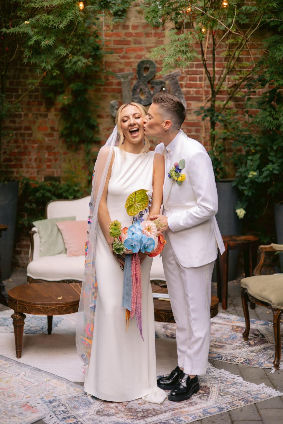 A bride in a white suit kissed a bride in a white dress on the cheek.