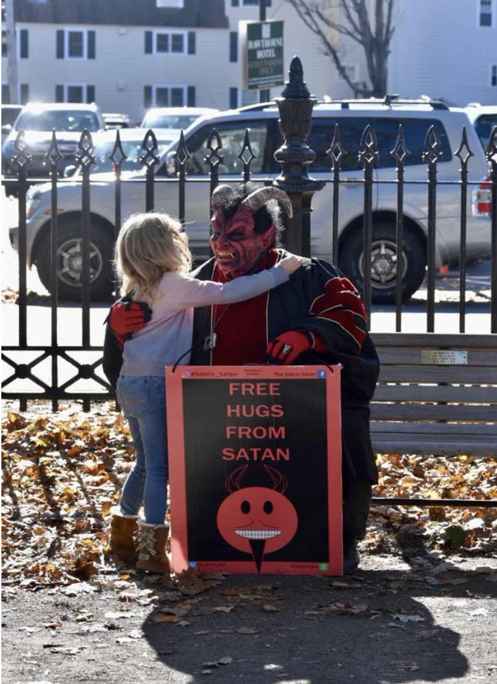 Little blonde girl hugging a man in a Satan costume in front of a sign "Free hugs from Satan"