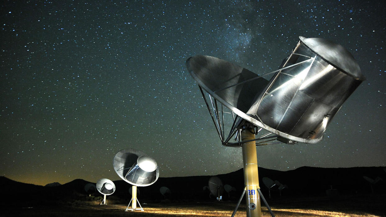  Spot-lit satellite dishes on an arid landscape point skyward ere the backdrop of a starry night. 