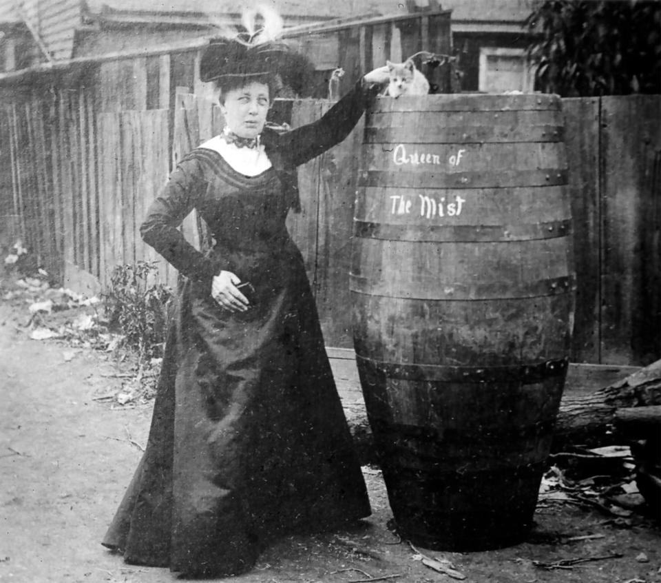 A woman in a high-necked, long dress with long sleeves and ornate hat standing next to a "Queen of the Mist" barrel