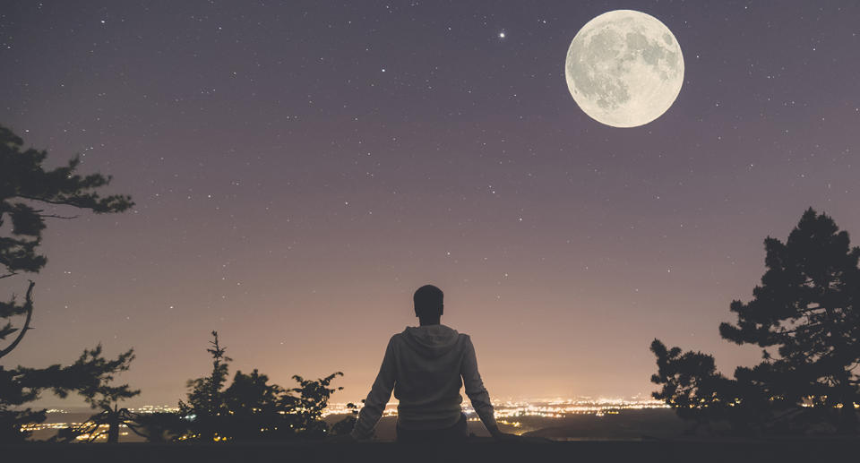 Photo shows a man sitting on a wall at night looking at the full moon.
