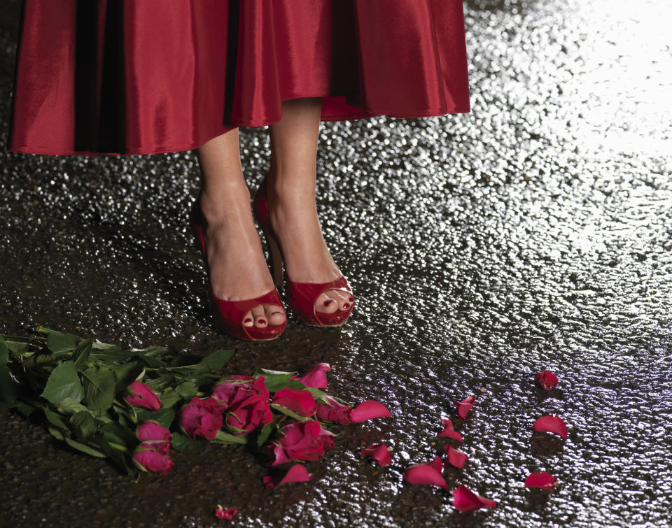 Close-up of a person's feet in red high heels standing on wet pavement next to a bouquet of red roses with petals scattered on the ground