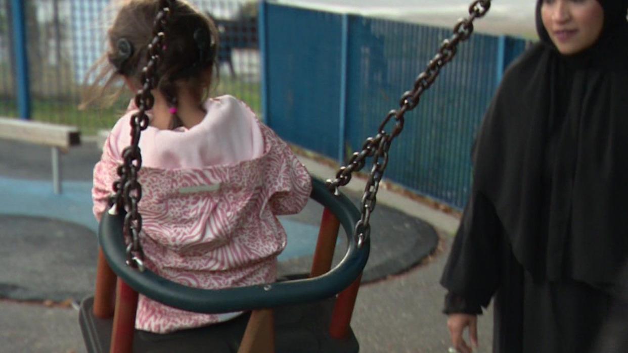 Aiyla, a child with a black cochlear implant and a pink coat, whose face is not visible, is pushed in an orange and black swing as her mother watches her