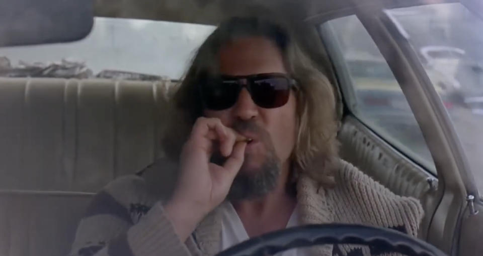 Jeff Bridges in "The Big Lebowski" is driving, wearing sunglasses, and smoking in a car. He has shoulder-length hair and a beard, and is wearing a casual cardigan