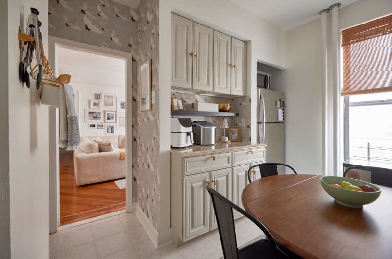 Fruit bowl on wood drop-leaf table in kitchen with beige cabinetry and wallpapered entry.