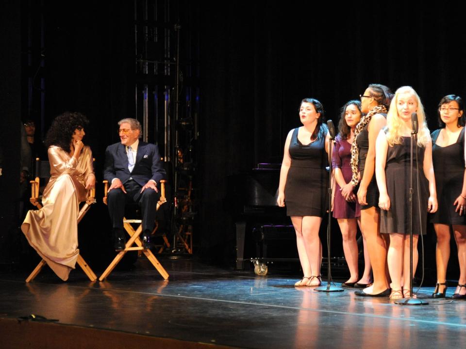 Gaga with black curly hair and a gold dress next to Bennett in a suit sitting on stage while students perform.