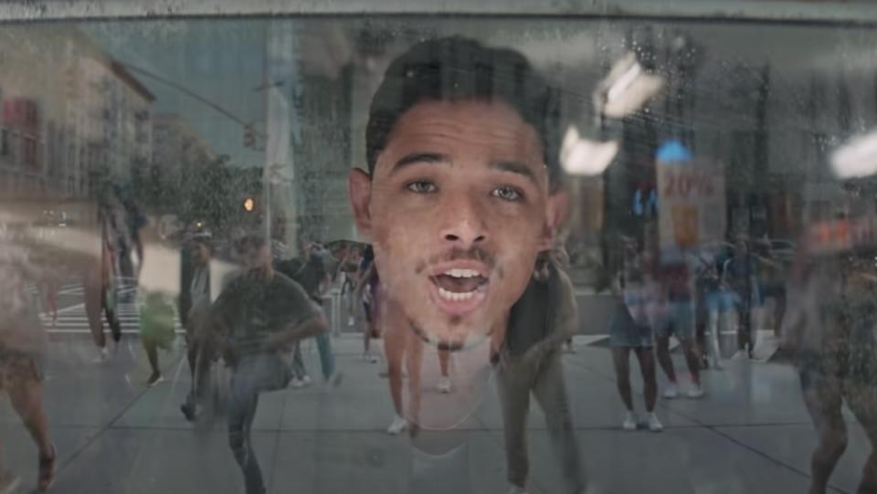 A man signs from inside a window as a street full of dancers is reflected in the glass