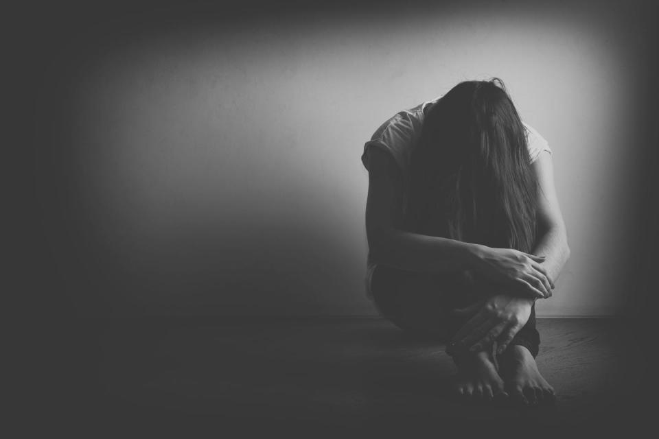 A black and white photo of a woman sitting on the floor with her knees drawn up and her head lowered