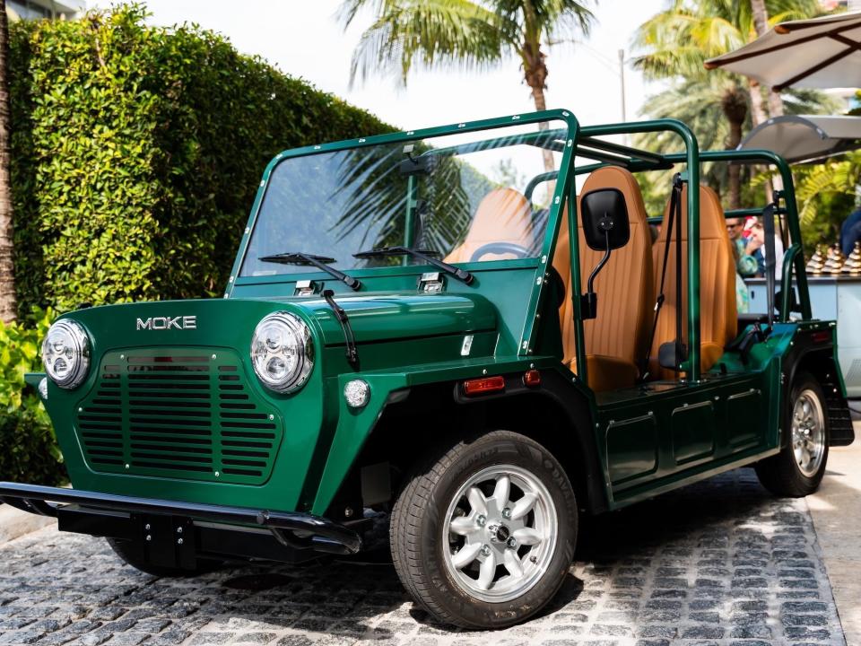 A green MOKE car with tan seats on a cobbled drive with palm trees in the background.