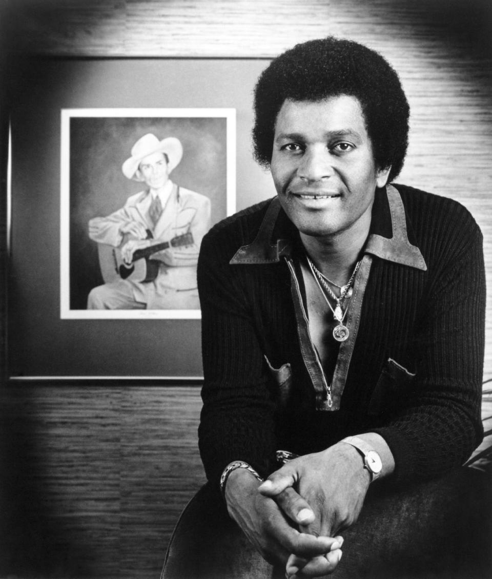 charley pride sitting in front of a portrait of hank williams and smiling for a photograph