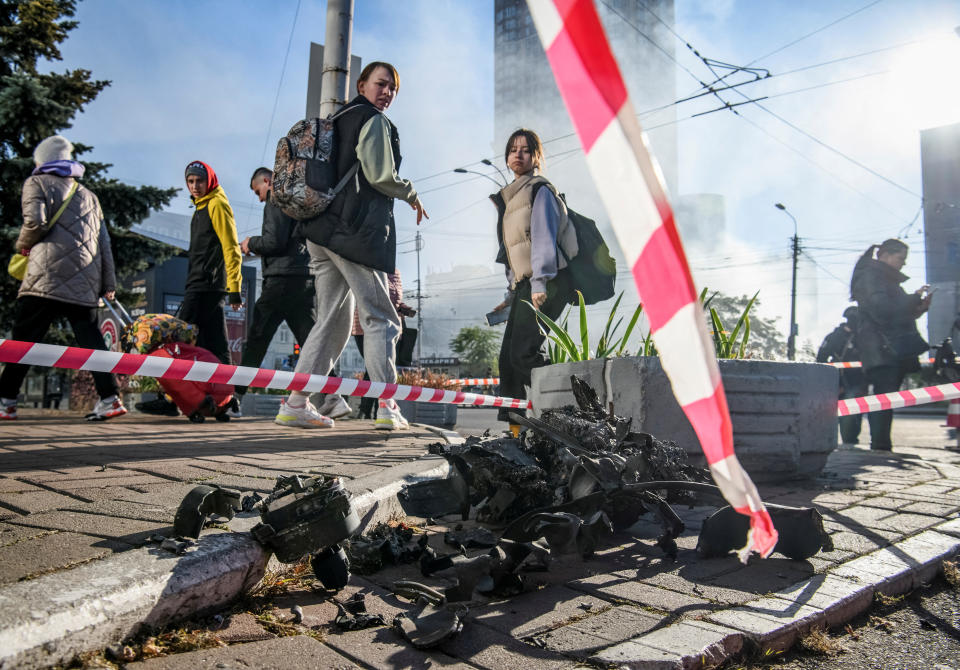 Two young women look back in disgust at a small pile of twisted metal on the cobblestones of a public street, surrounded by red-and-white striped emergency tape.