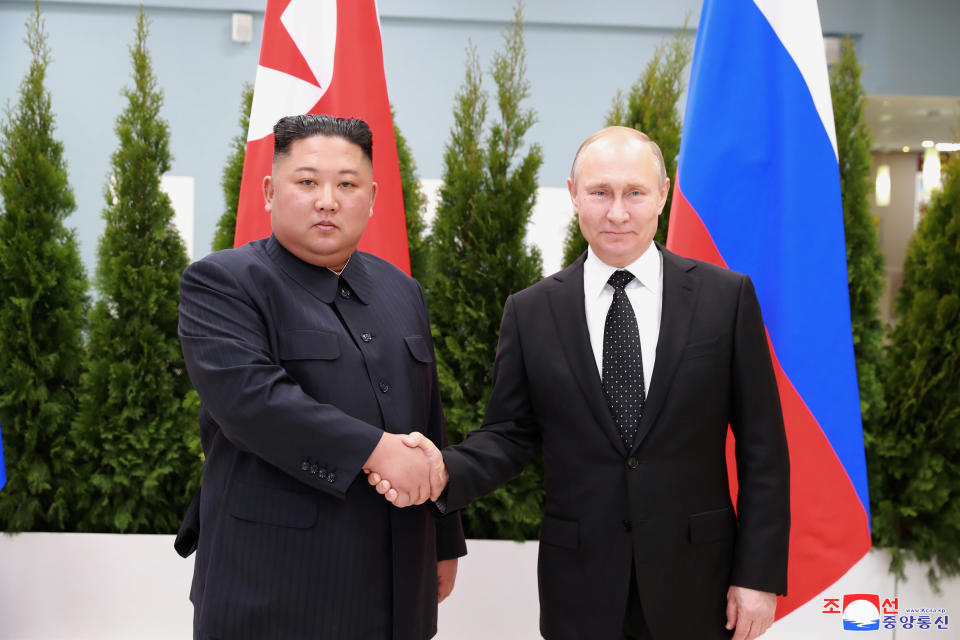 Kim Jong Un shakes hands with Vladimir Putin, both posing for the camera, in front of North Korean and Russian flags.